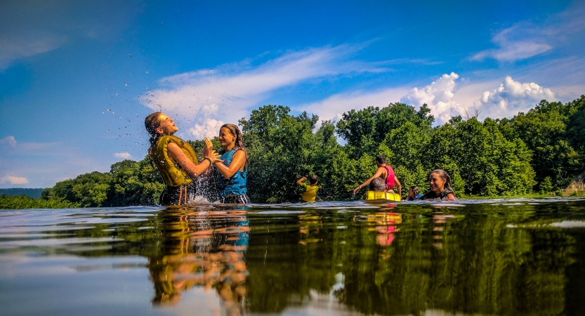 A group of young people play and splash in waist-deep water. They are wearing life jackets. There are green trees in the background and a blue sky above. 